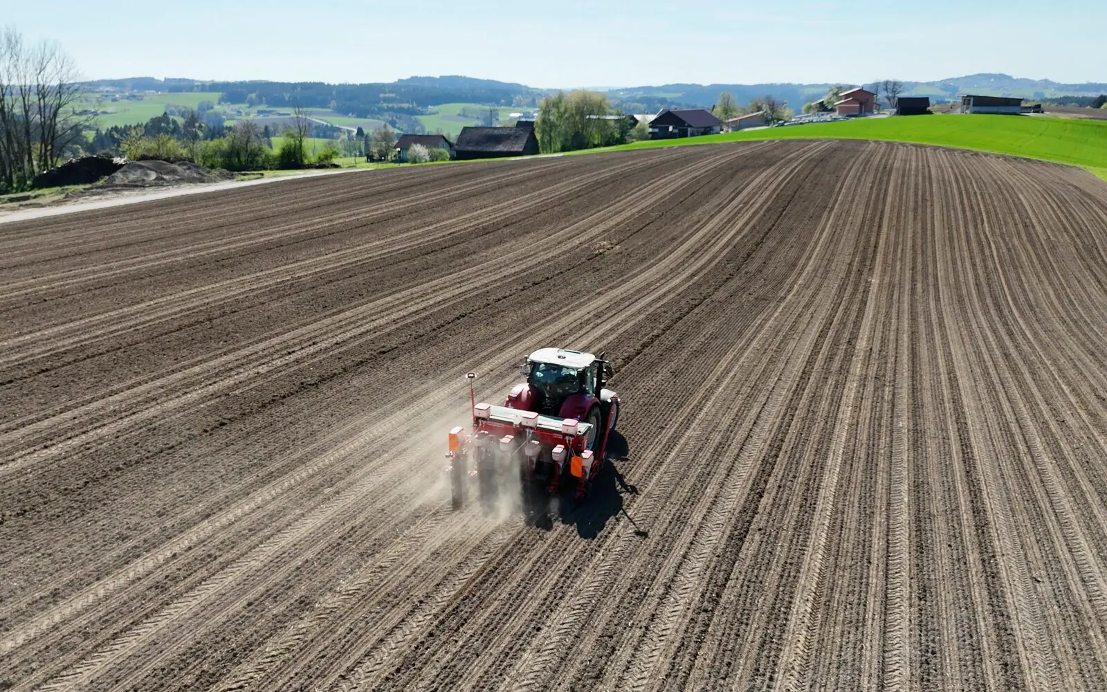 Traktor mit Sämaschine zieht gerade Spuren bis zum Horizont, dahinter Hügel und Höfe
